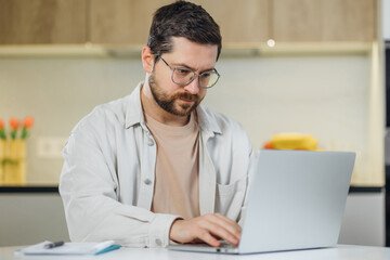 handsome young businessman in eyewear working with computer remotely, sitting at table in kitchen. Pleasant happy man searching information online.