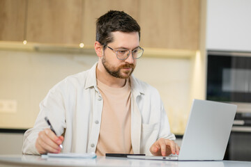 Happy handsome man with laptop and note pad at the kitchen table work remotely from home