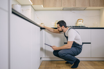 Leaning man in working clothes checking area under sink with clipboard at kitchen analyzing problem