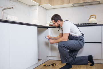 Serious repairman looking at contents of drainage system in kitchen and writing down info on clipboard holder