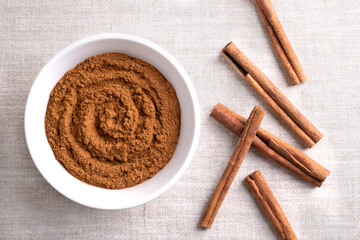 Cinnamon powder, in a white bowl on linen fabric. Raw bark of Indonesian cinnamon, Cinnamomum burmannii, ground and whole. Used mainly as aromatic condiment and flavouring additive in a wide variety.