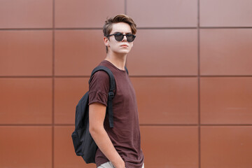 Handsome man with hairstyle with sunglasses in a fashion T-shirt with a backpack on a brown background on the street