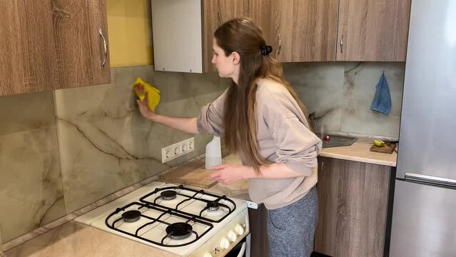 Woman cleaning kitchen backsplash with spray bottle and cloth in modern home kitchen, showcasing efficient house cleaning techniques