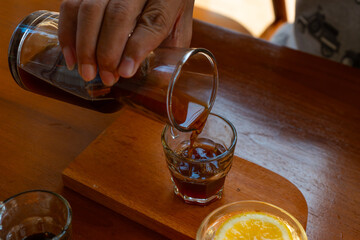 man's hand pouring v60 black coffee into small transparent glass, over wooden table surface, cafe environment