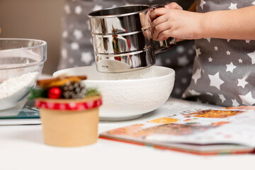 Metal cup sieve in the hands of a child, sifting flour, the process of making dough at Christmas.