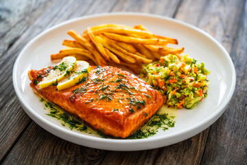 Fried salmon steak, French fries and vegetable salad served on white plate on wooden table

