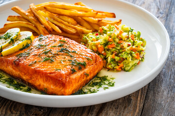 Fried salmon steak, French fries and vegetable salad served on white plate on wooden table
