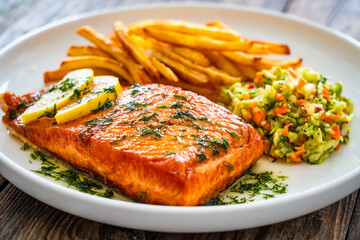 Fried salmon steak, French fries and vegetable salad served on white plate on wooden table
