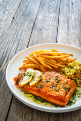 Fried salmon steak, French fries and vegetable salad served on white plate on wooden table
