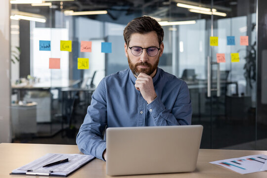 Photo of a thoughtful young man working concentratedly in the office at a laptop, sitting thoughtfully at a table against a transparent whiteboard and looking seriously at the screen