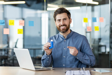 Portrait of a smiling young man sitting in the office at a table with a laptop, holding an asthma inhaler in his hand, pointing at it and looking at the camera