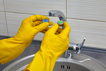 Plumber's hands in yellow rubber gloves change an old clogged kitchen sink faucet aerator to a new...