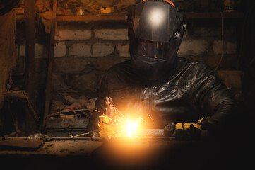 A welder in a protective mask for metal welding and protective gloves in a leather jacket performs welding work in a home garage. Sparks, hot flame from welding.