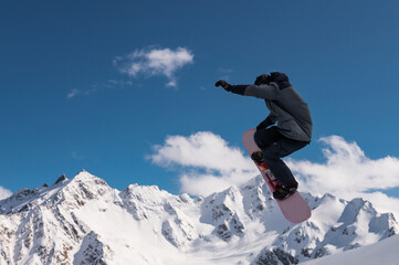 Portrait of a young man snowboarder jumping on a snowboard in sportswear, against the backdrop of high snow-capped mountains and sky