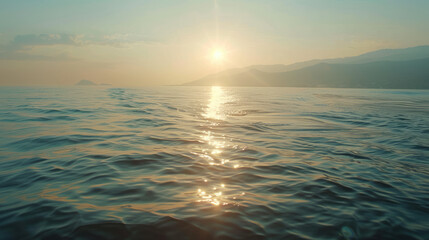 Fototapeta premium The sun is low over the sea, casting a glowing path on the water. Distant mountains are visible under a clear, pale sky. Waves gently ripple.