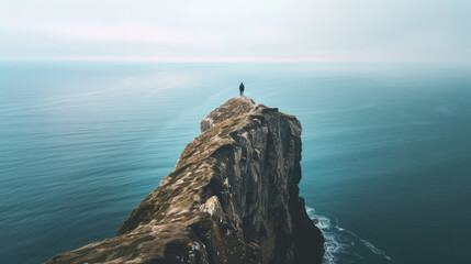 A person stands at the edge of a cliff, looking out over a vast, calm ocean under a cloudy sky. The cliff is high with rocks and some patches of grass.