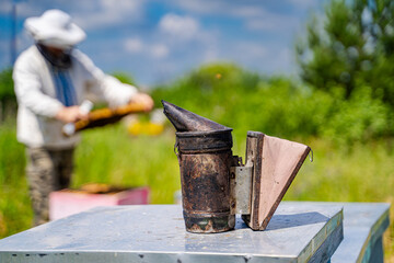 Bee Smoker On A Beehive. A bee smoker sits on top of a beehive, with a beekeeper working in the background.