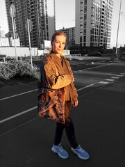 A beautiful young girl in a khaki bomber jacket stands against the background of high-rise buildings.