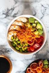 A poke bowl of food with a variety of vegetables including carrots, tomatoes, avocado and edamames and shrimps.
