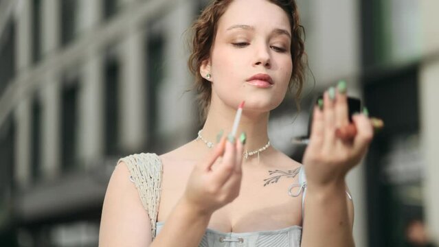 Pretty young caucasian woman stopped to check makeup and putting pomade lipstick for lips at city centre Charming female preparing for new day and smiling to mirror outdoors