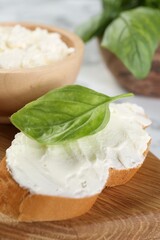 Piece of bread with cream cheese and basil leaf on white table, closeup