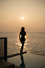 Fototapeta premium Woman in black swimsuit relaxing by infinity pool with ocean view during sunset.