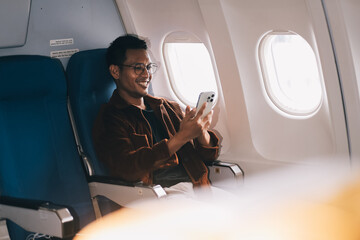 Happy curly man use phone and listens to music headphones on board airplane during flight.