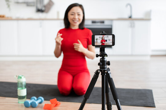 Woman in red activewear recording fitness video for online content, kneeling in home kitchen with exercise equipment and water bottle.