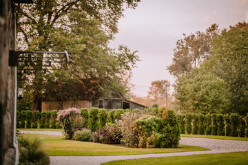 Blome, Latvia - September 11, 2023 - Beautiful garden with lush green bushes and trees, a gravel path, and a rustic wooden building in the background. Copy space...