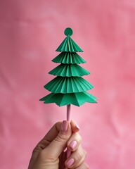 Minimalist Pink Background with Female Hand Holding Green Origami Christmas Tree Ornament