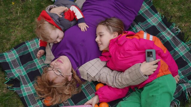 Slow motion. Top view. Mom and children lie on a plaid on the grass outdoors on a spring day. On one side, a mother hugs her teenage daughter, and on the other, she hugs and kisses her little son