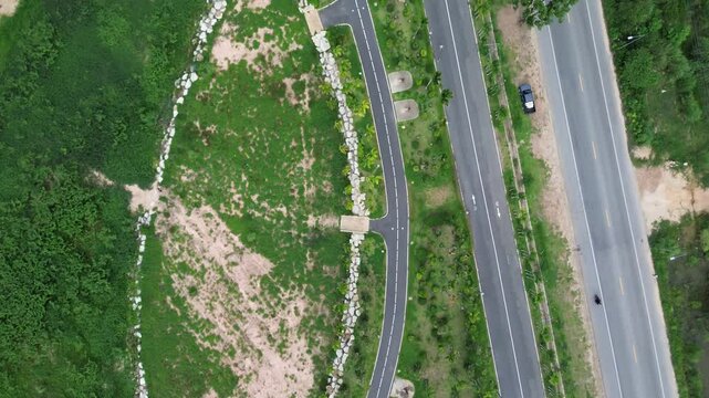 Drohnenaufnahme  eine gr&uuml;ne Landschaft in Thailand, durchzogen von einer Stra&szlig;e und einem asphaltierten Weg. Zwei Autos sind zu sehen, ein kleiner Bach und eine markante Steinbegrenzung trennen.