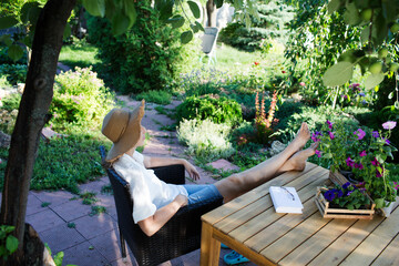happy middle aged woman   wearing a straw hat rests in a wicker chair in the garden near the house...
