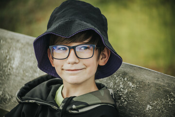 boy wearing glasses and a black bucket hat with a purple brim, sitting on a wooden bench outdoors. He wears a camouflage hoodie and has a playful expression on his face.