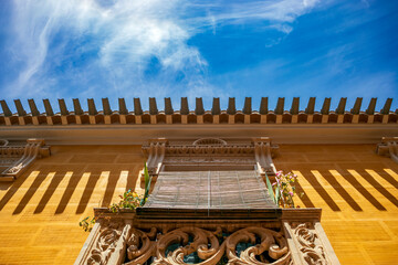 Detail of a yellow facade with a balcony with shutters and plants and the roof with gutters casting shadow on the facade in Pliego, Region of Murcia, Spain, in midday light