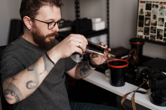A tattooed male photographer examines film negatives in a well-organized, contemporary home studio, surrounded by photography equipment. - Powered by Adobe