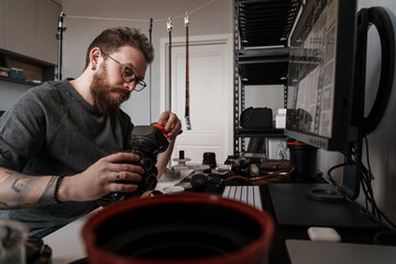 Concentrated male photographer with tattoos cleaning a camera lens in his home studio filled with various photographic equipment.