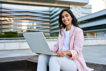 Smiling young middle eastern Israel businesswoman using laptop pc, looking at camera portrait. Indian or arabic woman freelancer in suit doing business analytics on computer, working remotely outdoors