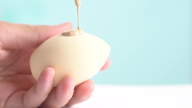 Make-up liquid beige foundation pouring on sponge, closeup. BB Cream dripping on beauty blender on blue white background. Macro Shot. Beautiful Makeup concept.