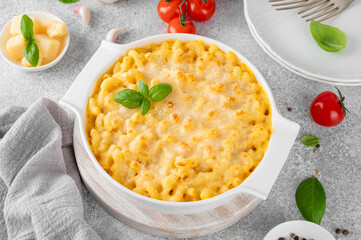 Mac and cheese in a white baking dish on a wooden board on a gray concrete background. American cuisine, comfort food. Copy space.