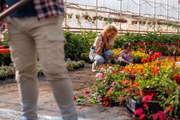 A mother and daughter kneeling and smiling while gardening in a greenhouse, surrounded by vibrant...