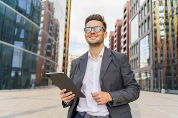 Fototapeta premium A cheerful businessman in a suit holds a tablet, standing confidently amid modern urban buildings.