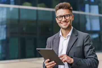 A smiling businessman in a suit uses a tablet, standing confidently in a modern urban environment.