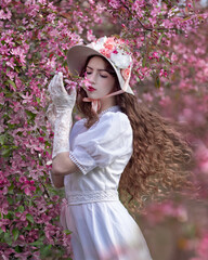 Portrait of a girl in a white dress and hat against the background of a blooming pink apple tree with flowing hair