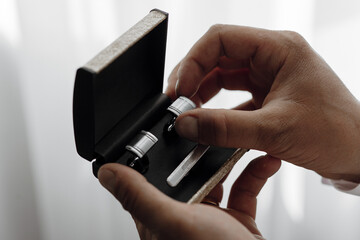 A man is holding a pair of cufflinks in a box. The cufflinks are silver and have a gold trim. The man is wearing a white shirt and he is getting ready for a formal event