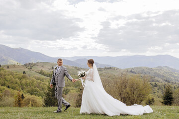 A bride and groom are walking on a grassy hillside, with the bride wearing a long white dress. The scene is serene and picturesque