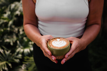 Woman in white top holding cup cappuccino on plant background
