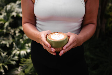 Woman in white top holding green mug with cappuccino on plant background