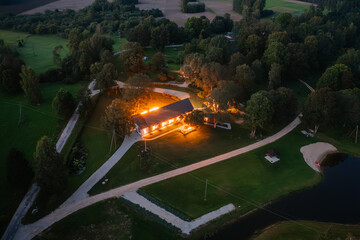 Blome, Latvia - September 11, 2023 - Aerial view of a warmly lit house at dusk, surrounded by trees, pathways, and a pond, creating a serene and cozy atmosphere.