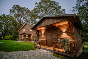 Blome, Latvia - September 11, 2023 - Two wooden cabins with illuminated exteriors, surrounded by greenery and paved walkways, nestled under large trees in a serene setting.